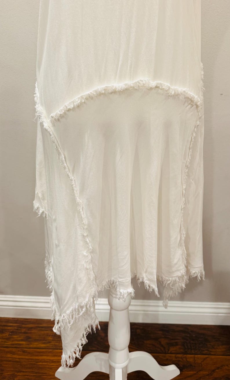 Puede incluir: Vestido blanco asim&eacute;trico con bordes deshilachados. El vestido se exhibe sobre un pedestal blanco. La tela parece ligera y fluida, con un dise&ntilde;o en capas y un ribete texturizado y con flecos.