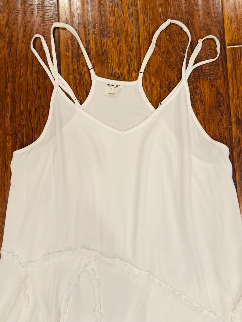Puede incluir: Un camis&oacute;n blanco con tirantes finos y cuello en V. La parte superior tiene un dise&ntilde;o fluido con un dobladillo con volantes. La etiqueta dice "INTIMATELY". La prenda est&aacute; hecha de una tela ligera.