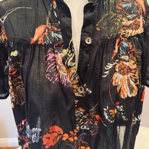 May include: Black button-down shirt with a vibrant floral pattern in shades of orange, pink, and blue. The short-sleeved shirt features a mandarin collar and a semi-sheer fabric, offering a lightweight and airy feel. The floral design is spread across the shirt.