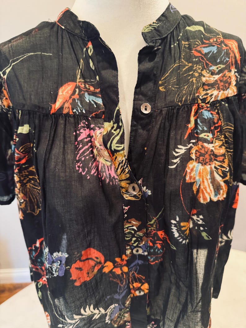 May include: Black button-down shirt with a vibrant floral pattern in shades of orange, pink, and blue. The short-sleeved shirt features a mandarin collar and a semi-sheer fabric, offering a lightweight and airy feel. The floral design is spread across the shirt.