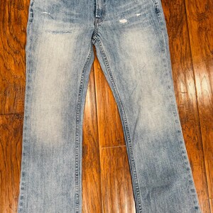 Puede incluir: Vaqueros de mezclilla azul claro con un lavado descolorido y detalles desgastados. Los vaqueros tienen un dise&ntilde;o cl&aacute;sico de cinco bolsillos, cierre de bot&oacute;n y cremallera, y un dobladillo sin rematar deshilachado. Los vaqueros est&aacute;n sobre una superficie de madera.