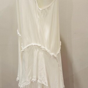 Puede incluir: Vestido blanco sin mangas con falda de capas y flecos. El vestido tiene un dise&ntilde;o simple y elegante con una silueta fluida. El vestido se exhibe en un maniqu&iacute;, mostrando su forma y textura. El vestido est&aacute; hecho de una tela suave y ligera.