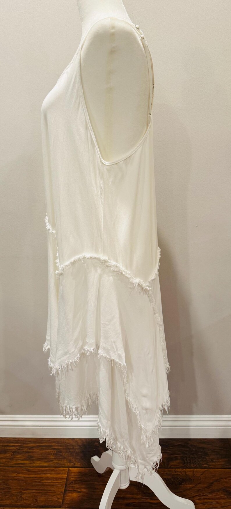 Puede incluir: Vestido blanco sin mangas con falda de capas y flecos. El vestido tiene un dise&ntilde;o simple y elegante con una silueta fluida. El vestido se exhibe en un maniqu&iacute;, mostrando su forma y textura. El vestido est&aacute; hecho de una tela suave y ligera.