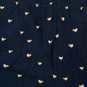 May include: Close-up of a dark navy fabric with a repeating pattern of small hearts. Each heart is a combination of white and yellow, creating a playful and cheerful design. The fabric has a textured appearance.