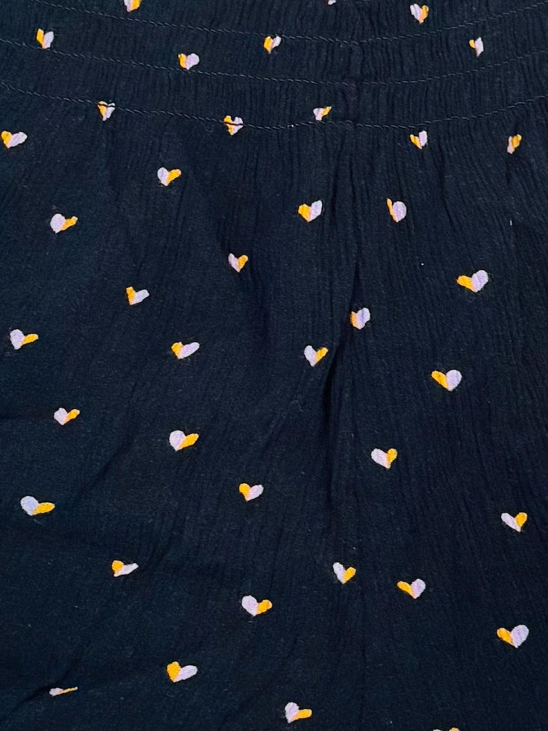 May include: Close-up of a dark navy fabric with a repeating pattern of small hearts. Each heart is a combination of white and yellow, creating a playful and cheerful design. The fabric has a textured appearance.