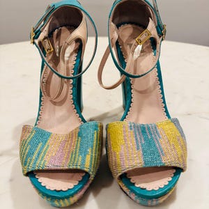 May include: Pair of teal and multi-colored open-toe platform sandals. The sandals feature a woven fabric design in shades of blue, yellow, and pink. They have ankle straps with gold buckles and scalloped edges. The heels are approximately 4 inches.