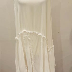 Puede incluir: Vestido blanco y fluido con tirantes finos y cuello en V. El vestido presenta un dise&ntilde;o escalonado con bordes deshilachados, lo que le da una est&eacute;tica bohemia. El vestido se exhibe en un maniqu&iacute;.