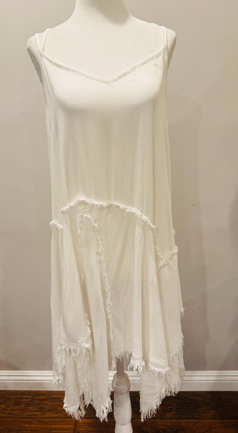 Puede incluir: Vestido blanco y fluido con tirantes finos y cuello en V. El vestido presenta un dise&ntilde;o escalonado con bordes deshilachados, lo que le da una est&eacute;tica bohemia. El vestido se exhibe en un maniqu&iacute;.