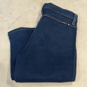 May include: Dark blue denim jeans folded neatly. The jeans have a classic five-pocket design, with a button and zipper closure. The stitching is a contrasting tan color. The brand name " Frankie B." is visible on the back waistband.