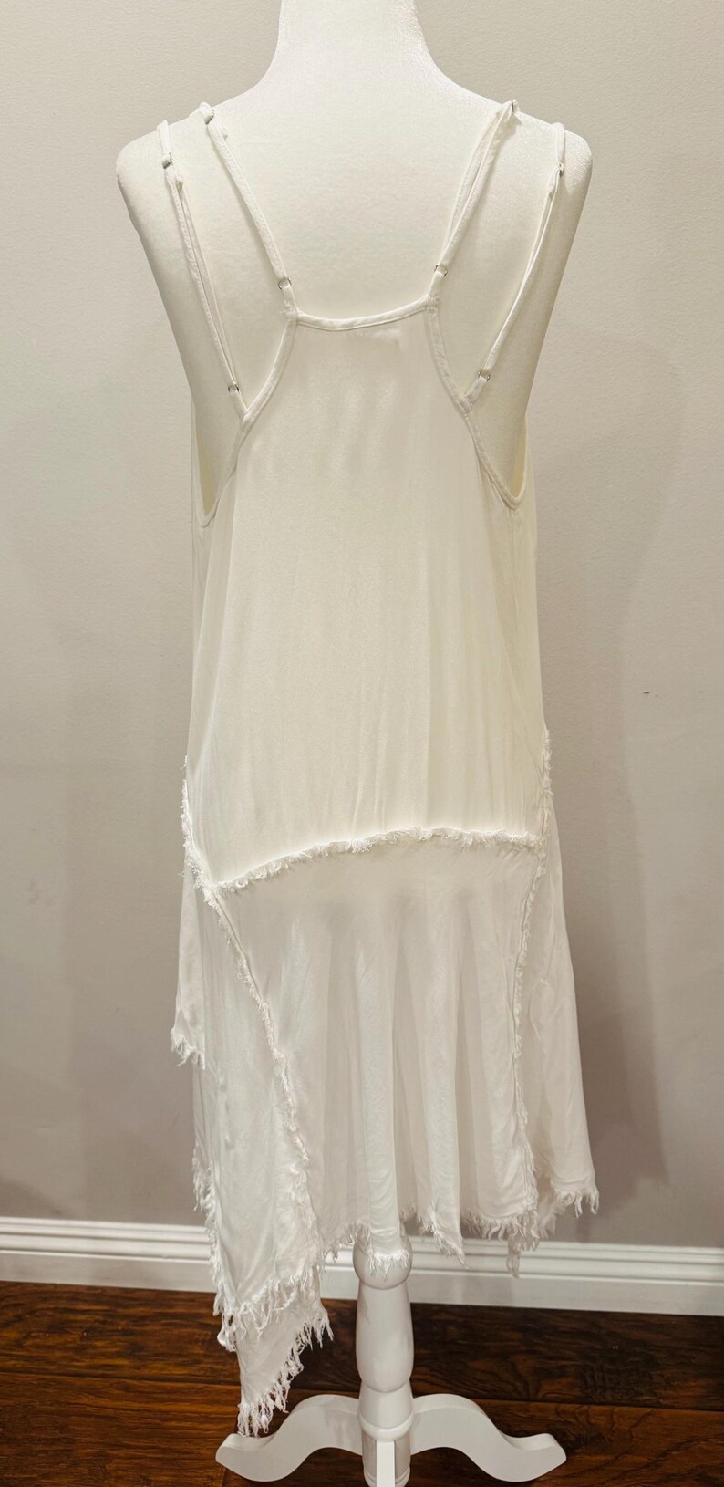 Puede incluir: Vestido blanco sin mangas con tirantes finos y dobladillo asim&eacute;trico. El vestido presenta un dise&ntilde;o en capas con bordes sin rematar, lo que le da una est&eacute;tica bohemia o vintage. El vestido se exhibe en un maniqu&iacute;.