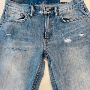 Puede incluir: Vaqueros de mezclilla azul claro con detalles desgastados. Los vaqueros tienen cierre de bot&oacute;n, bolsillos delanteros y traseros y un lavado descolorido. La marca "ALLSAINTS" es visible en una etiqueta. Los vaqueros tienen detalles rasgados en la parte delantera.