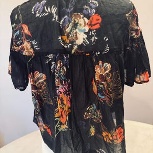 May include: Black short-sleeved blouse with a floral pattern in shades of orange, brown, purple, and white. The blouse has a collared neckline and a slightly ruffled hem. The fabric appears to be lightweight and semi-sheer.