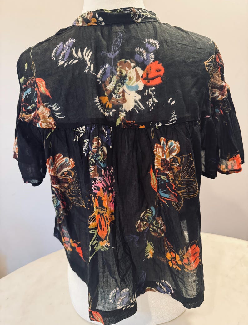 May include: Black short-sleeved blouse with a floral pattern in shades of orange, brown, purple, and white. The blouse has a collared neckline and a slightly ruffled hem. The fabric appears to be lightweight and semi-sheer.