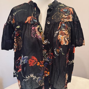 May include: Black button-down shirt with a floral pattern in shades of orange, pink, blue, and yellow. The short-sleeved blouse has a mandarin collar and a semi-sheer appearance. The floral print is scattered across the fabric.