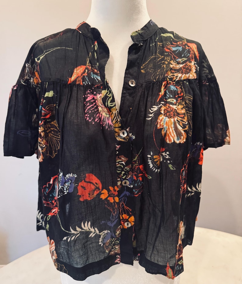 May include: Black button-down shirt with a floral pattern in shades of orange, pink, blue, and yellow. The short-sleeved blouse has a mandarin collar and a semi-sheer appearance. The floral print is scattered across the fabric.