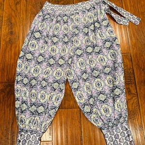 K&ouml;nnte beinhalten: Gemusterte Hose mit einem hellvioletten, gelben und marineblauen Blumenmuster. Die Hose hat eine geraffte Taille mit Bindeb&auml;ndern und Manschetten mit Kn&ouml;pfen. Der Stoff hat ein weiches, flie&szlig;endes Aussehen.