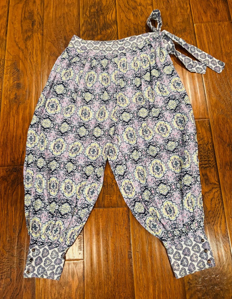 K&ouml;nnte beinhalten: Gemusterte Hose mit einem hellvioletten, gelben und marineblauen Blumenmuster. Die Hose hat eine geraffte Taille mit Bindeb&auml;ndern und Manschetten mit Kn&ouml;pfen. Der Stoff hat ein weiches, flie&szlig;endes Aussehen.