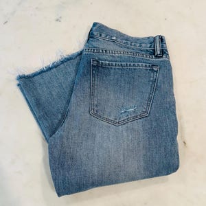Puede incluir: Pantalones cortos vaqueros azul claro doblados con dobladillo deshilachado y bolsillo trasero. Los pantalones cortos tienen un aspecto desgastado con un peque&ntilde;o desgarro cerca del bolsillo trasero. Los pantalones cortos est&aacute;n doblados cuidadosamente sobre una superficie blanca.