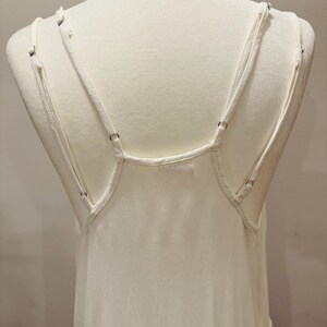 Puede incluir: Camisola de color marfil con tirantes finos ajustables. Las correas est&aacute;n unidas a la espalda con peque&ntilde;os anillos met&aacute;licos. La tela parece ser un material suave y fluido, y la prenda se exhibe en un maniqu&iacute;.