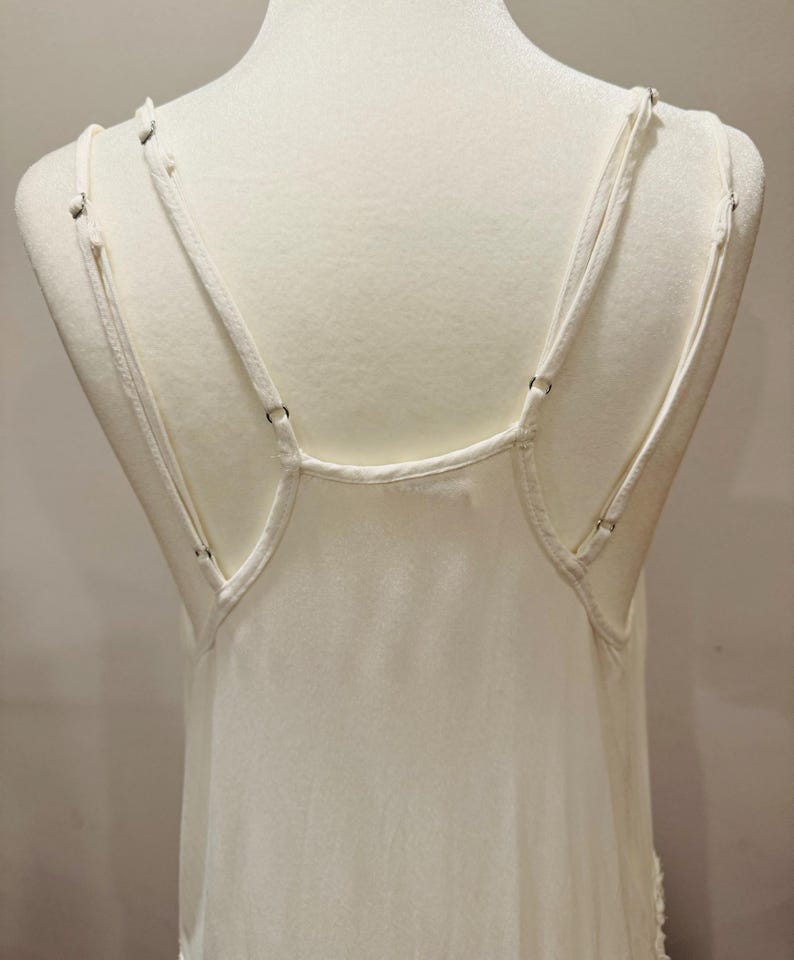Puede incluir: Camisola de color marfil con tirantes finos ajustables. Las correas est&aacute;n unidas a la espalda con peque&ntilde;os anillos met&aacute;licos. La tela parece ser un material suave y fluido, y la prenda se exhibe en un maniqu&iacute;.
