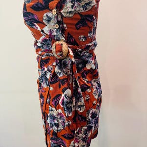 K&ouml;nnte beinhalten: Ein tr&auml;gerloses Kleid mit floralem Muster in Orange-, Wei&szlig;-, Lila- und Marineblaut&ouml;nen. Das Kleid hat Holzkn&ouml;pfe, eine Holzschlie&szlig;e und ein Wickeldesign. Der Stoff scheint leicht zu sein.
