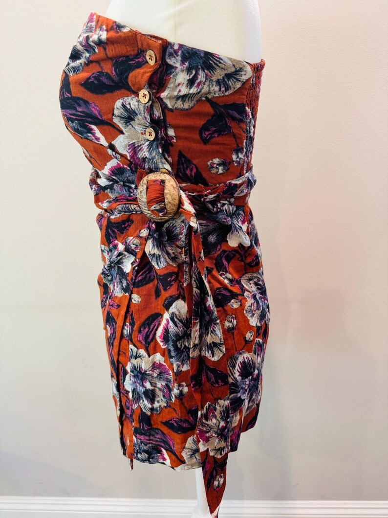 K&ouml;nnte beinhalten: Ein tr&auml;gerloses Kleid mit floralem Muster in Orange-, Wei&szlig;-, Lila- und Marineblaut&ouml;nen. Das Kleid hat Holzkn&ouml;pfe, eine Holzschlie&szlig;e und ein Wickeldesign. Der Stoff scheint leicht zu sein.