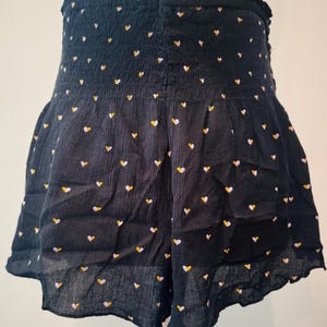 May include: Black shorts with a high, elasticized waist and a ruffled hem. The shorts are covered in a pattern of small hearts in yellow, white, and pink. The fabric has a textured appearance.
