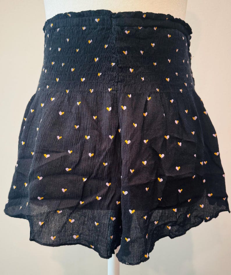 May include: Black shorts with a high, elasticized waist and a ruffled hem. The shorts are covered in a pattern of small hearts in yellow, white, and pink. The fabric has a textured appearance.