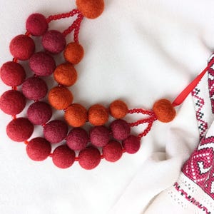 Puede incluir: Un collar hecho a mano con bolas de fieltro en tonos rojos y naranjas. El collar está ensartado en un cordón rojo con pequeñas cuentas rojas. El collar se exhibe sobre un fondo blanco con una porción de tela blanca con bordados rojos.
