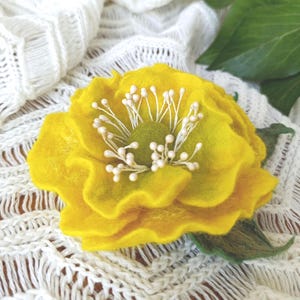 Puede incluir: Una flor de fieltro amarillo hecha a mano con un centro verde y estambres de cuentas blancas. La flor tiene múltiples pétalos rizados y está colocada sobre un fondo blanco texturizado. Se ven hojas de fieltro verdes.