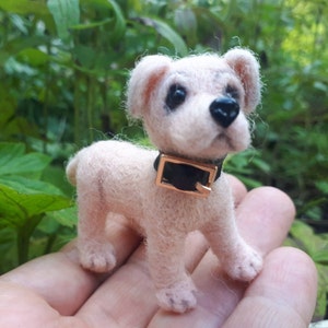 Puede incluir: Una pequeña figura de perro hecha con lana fieltrada con aguja en color rosa claro. El perro tiene ojos oscuros y brillantes y una nariz negra. Lleva un collar negro con una hebilla rectangular de color dorado metálico y textura visible.