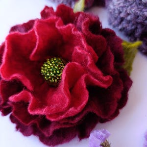 Puede incluir: Una flor de fieltro rojo oscuro hecha a mano con un centro verde con cuentas. La flor tiene pétalos rizados y una hoja verde. El fondo incluye un artículo de punto morado y flores moradas.