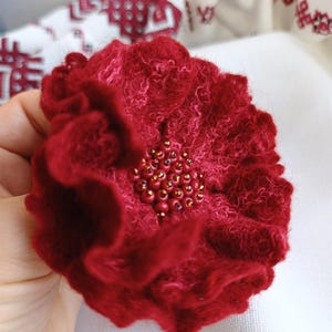Puede incluir: Una flor de fieltro rojo oscuro hecha a mano, con un centro de cuentas. La flor tiene múltiples pétalos rizados y un aspecto texturizado. El centro está adornado con pequeñas cuentas redondas de color marrón dorado. Probablemente un broche o un accesorio decorativo.