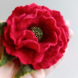 Puede incluir: Una flor roja de fieltro hecha a mano con un centro de cuentas verdes y hojas de fieltro verdes. La flor tiene múltiples capas de pétalos, creando una apariencia texturizada. El fieltro es de color rojo vibrante.