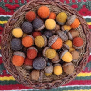 Puede incluir: Una cesta de mimbre llena de bellotas de fieltro de colores. Las bellotas tienen tapas marrones y cuerpos de fieltro en tonos naranja, amarillo y morado. La cesta está sobre un textil tejido colorido.