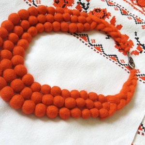 Puede incluir: Un collar de bolas de fieltro naranja. El collar presenta tres hilos de bolas de fieltro, conectados por una sección trenzada y un cierre plateado. Se muestra sobre una tela blanca con bordados florales naranjas.