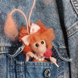 May include: A handmade felt poodle brooch with orange fur, a pink bow, and a pearl necklace, holding a red lollipop. The poodle is nestled in the pocket of a blue denim jacket.