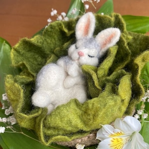 May include: A small, white and grey felt bunny rabbit is sleeping in a green felt nest. The nest is surrounded by green leaves and white flowers.