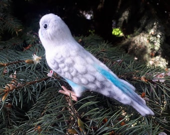 Puede incluir: Un pájaro de fieltro hecho a mano, principalmente blanco con acentos grises y azules, posado en una rama de pino verde. El pájaro tiene una apariencia texturizada detallada, con un pico pequeño y un ojo oscuro. Las plumas de la cola y las alas son de color azul brillante.