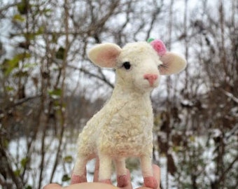 Pode incluir: Uma figura de cordeiro artesanal, creme, com nariz e cascos rosa, e uma pequena flor rosa na cabeça. O cordeiro tem orelhas grandes e caídas e está em pé. O fundo é uma cena exterior desfocada com árvores nuas.