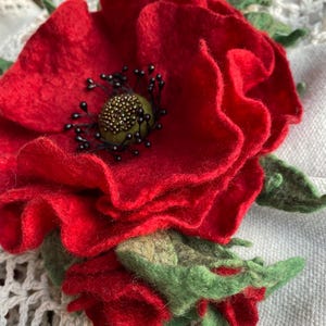 Puede incluir: Una amapola roja de fieltro hecha a mano, con hojas verdes y un centro detallado. La amapola tiene pétalos en capas y un aspecto texturizado. El centro es una mezcla de cuentas doradas y negras. Un hermoso accesorio floral.