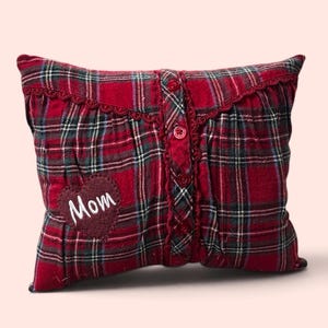 May include: A rectangular throw pillow featuring a red, green, and white plaid pattern. The pillow has a vertical button placket with four red buttons and a decorative lace trim. A dark red heart-shaped patch with the white embroidered text Mom.