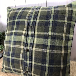 May include: Square pillow with a green, navy, and cream plaid pattern. The pillow is made from a button-down shirt, with the buttons and pocket visible. The pillow is a decorative item for a bed or couch.