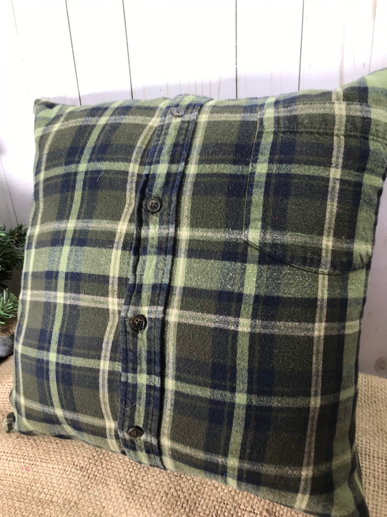 May include: Square pillow with a green, navy, and cream plaid pattern. The pillow is made from a button-down shirt, with the buttons and pocket visible. The pillow is a decorative item for a bed or couch.