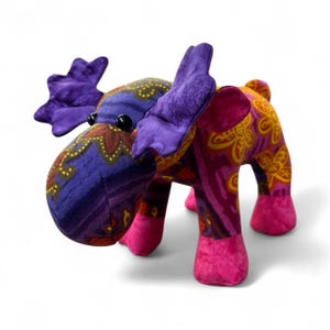 May include: A plush moose toy with a vibrant, patterned design. The moose features purple antlers, a purple and blue head with a floral pattern, and a body in shades of pink and yellow. The hooves are bright pink, and the eyes are black.