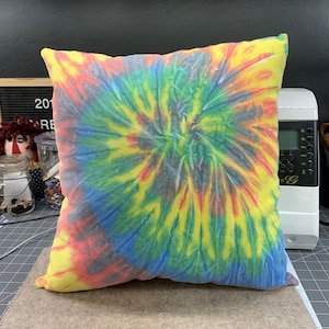 May include: A square tie-dye pillow with a spiral pattern in yellow, red, blue, green, and gray. The pillow is on a brown surface. The pillow is a home decor item.