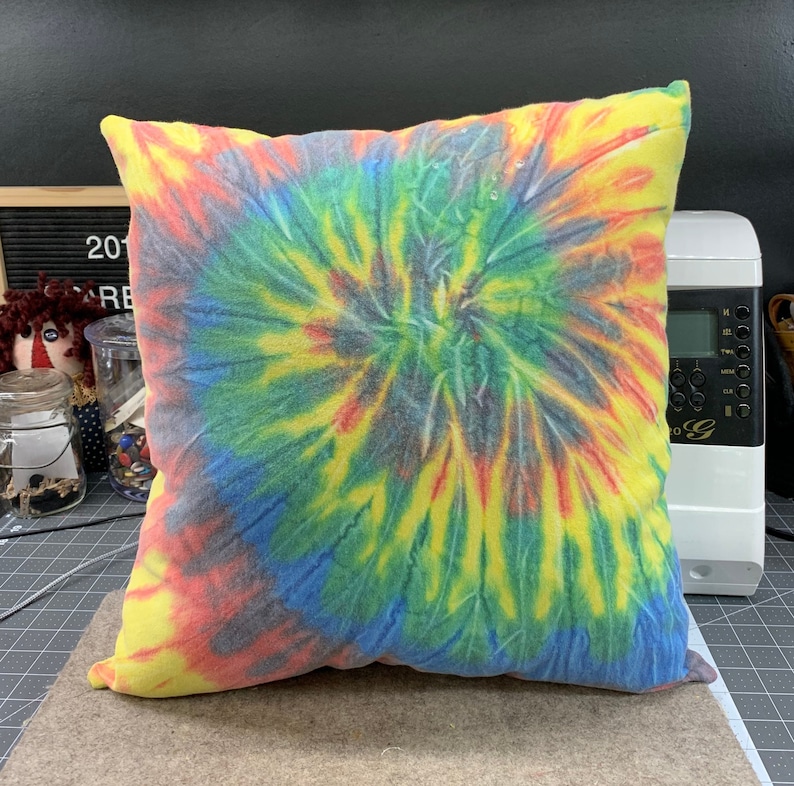 May include: A square tie-dye pillow with a spiral pattern in yellow, red, blue, green, and gray. The pillow is on a brown surface. The pillow is a home decor item.