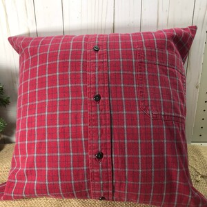 May include: A square pillow made from a red plaid shirt. The pillow features a button-down front with black buttons and a pocket on the right side. The plaid pattern includes red, black, and light blue lines.