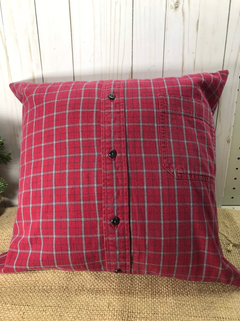 May include: A square pillow made from a red plaid shirt. The pillow features a button-down front with black buttons and a pocket on the right side. The plaid pattern includes red, black, and light blue lines.