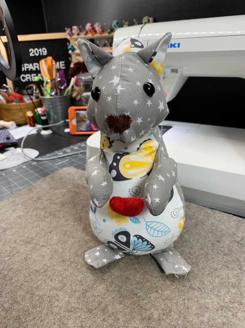 May include: A grey and white fabric squirrel holding a red heart in its paw. The squirrel has a star pattern on its head and a butterfly pattern on its body.